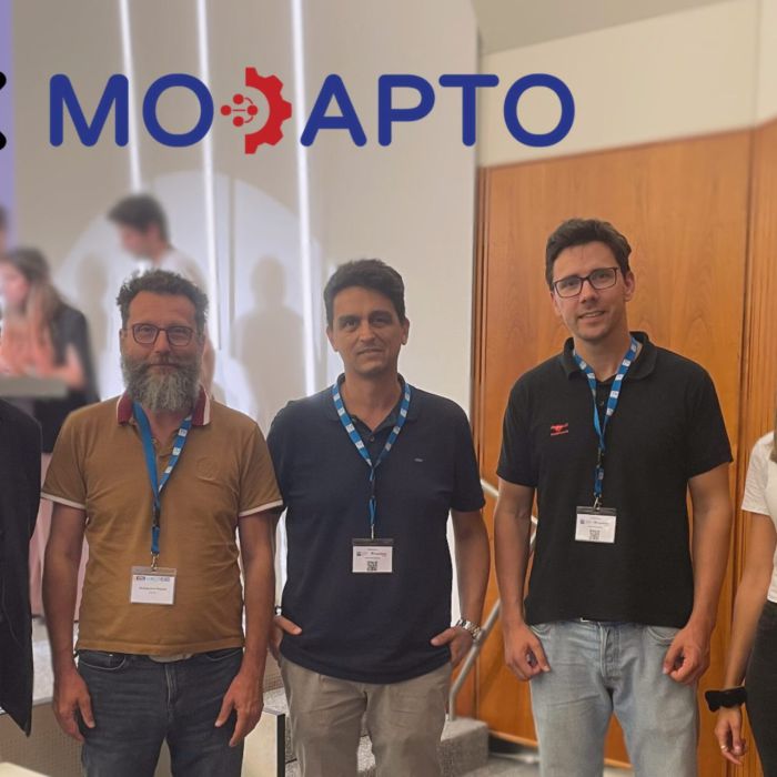 Group of five Manufacturing researchers of the AMiMO Cluster: one woman and four men (three of them wear glasses). They all have blue ticket holders around their necks and are smiling into the camera. In the background there are three people standing on a podium but they are blurred out. At the top left there is the MARS logo and the MODAPTO logo, both projects funded by the EU under the same call category. They are also members of the AMiMO Cluster.